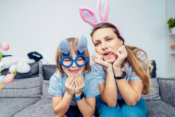 portrety szczęśliwej, uśmiechniętej matki rasy kaukaskiej i jej syna w uszach królika bawiących się w domu w wielkanoc. rodzina siedzi na kanapie w domu, bawi się w małpę, szaleje. tradycje wielkanocne. selektywna ostrość - easter rabbit sofa women zdjęcia i obrazy z banku zdjęć