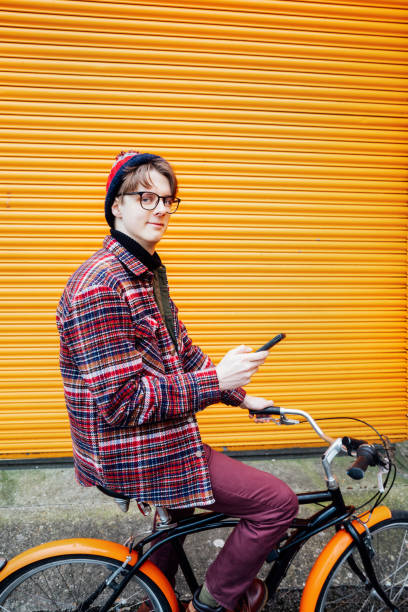 istock Young hipster man using mobile phone while sitting on city bicycle near orange wall. Student using 5g network,rests after riding. Eco friendly transport for carbon footprint. Sustainable lifestyle 2063221377