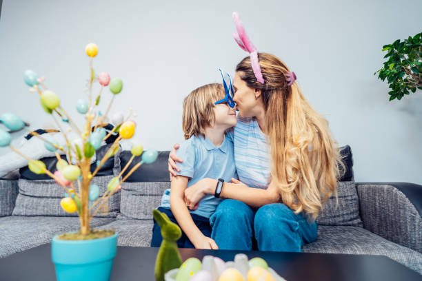 szczęśliwa uśmiechnięta kaukaska mama i jej syn w uszach królika bawią się w domu w wielkanoc. rodzina siedzi na kanapie w domu i przytula się, matka całuje syna. rodzinne tradycje wielkanocne. selektywna ostrość - easter rabbit sofa women zdjęcia i obrazy z banku zdjęć