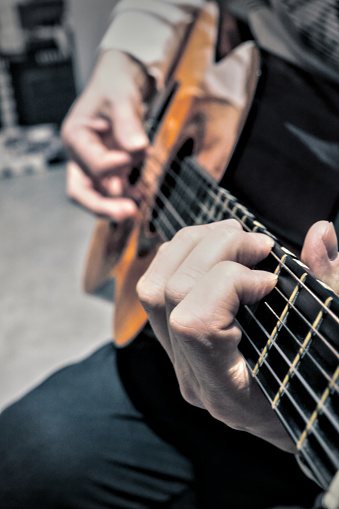 Close-up on the hands of a man playing classical guitar, seated on a leather armchair. The hand strumming the strings is out of focus.