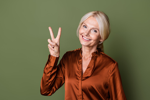 Photo portrait of lovely senior lady show v-sign nonverbal dressed stylish silk brown garment isolated on khaki color background.