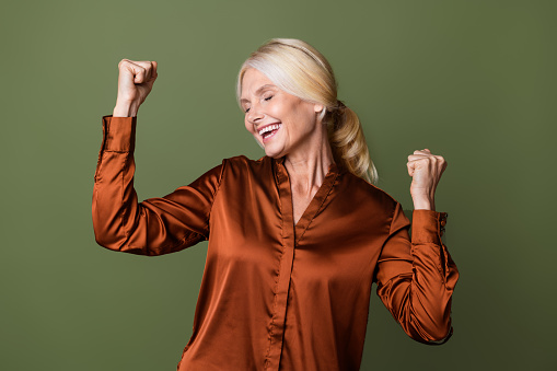 Photo portrait of attractive pensioner woman raise fists celebrate dressed stylish silk brown clothes isolated on khaki color background.