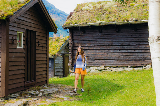 Smiling female explorer with long hair, wearing stripe shirt and orange shorts running between old viking houses with grass on roof on the fresh green meadow in Loen, Western Norway, Scandinavia