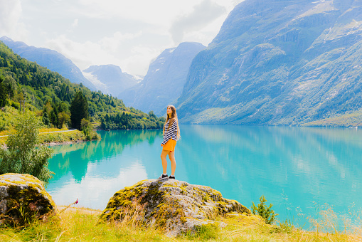 FroSident View of happy female traveler in orange shorts staying on the rock with scenic viewpoint of glacial mountain lake Lovatnet in Western Norway, Scandinavia