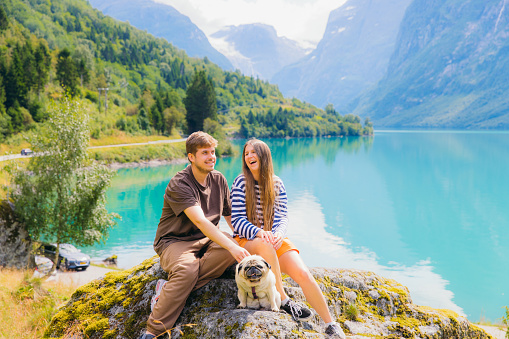 Smiling woman and man traveler with their cute pug exploring Scandinavia during summertime - they sitting on the rock with scenic view of glacial lake Lovatnet and beautiful mountain peaks in Stryn
