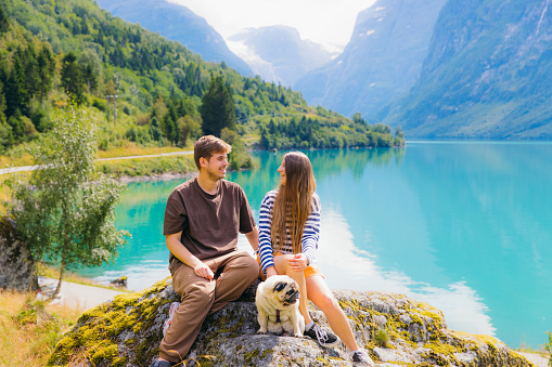 Smiling woman and man traveler with their cute pug exploring Norway during summertime - they sitting on the rock with scenic view of glacial lake Lovatnet and beautiful mountain peaks in Stryn