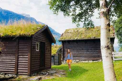 Smiling female explorer with long hair, wearing stripe shirt and orange shorts running between old viking houses with grass on roof on the fresh green meadow in Loen, Western Norway, Scandinavia