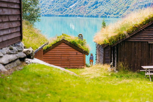 Smiling female explorer with long hair, wearing stripe shirt and orange shorts staying between old viking houses on the fresh green meadow looking at glacial lake Lovatnet in Loen, Western Norway, Scandinavia