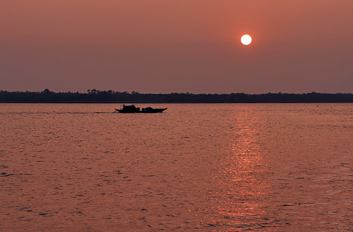 silhouette of a small fishing boat rowing through vast waterscape of tidal river at Sundarbans biosphere reserve. The last rays of sun creating magical atmosphere with golden warm lights.\nSundarbans is a popular place for nature lovers and part of growing ecotourism boom.