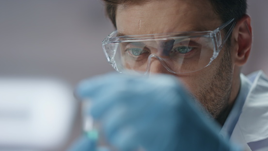 Clinic worker analyzing sample experimental substance in test tube close up. Serious scientist in gloves glasses entering research data in lab computer. Smart biochemist examining coronavirus vaccine.