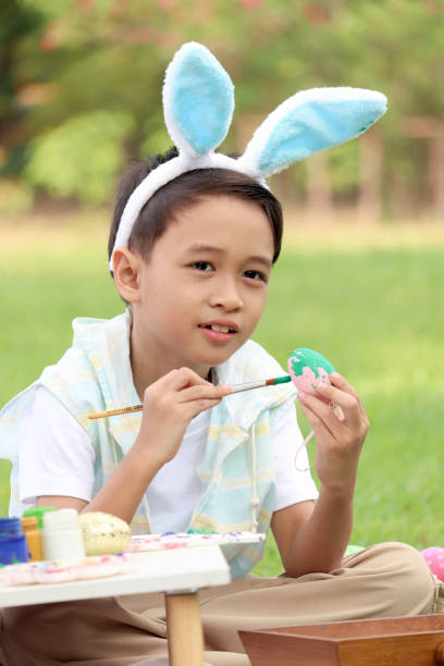 ウサギの耳を持つかわいいアジアの男の子は、自然の庭の緑の草原に座って絵筆で卵を描いています。イースター休暇を屋外で祝う子供。幸せな子供はイースターに公園で楽しんでいます - easter easter bunny fun humor ストックフォトと画像