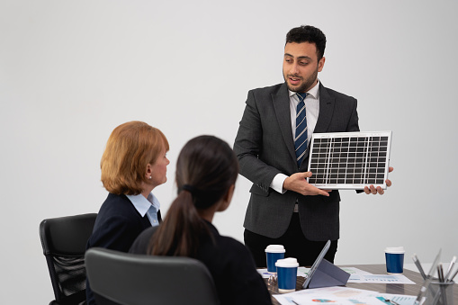 A confident businessman presents a solar panel to colleagues, demonstrating renewable energy solutions in a corporate environment.