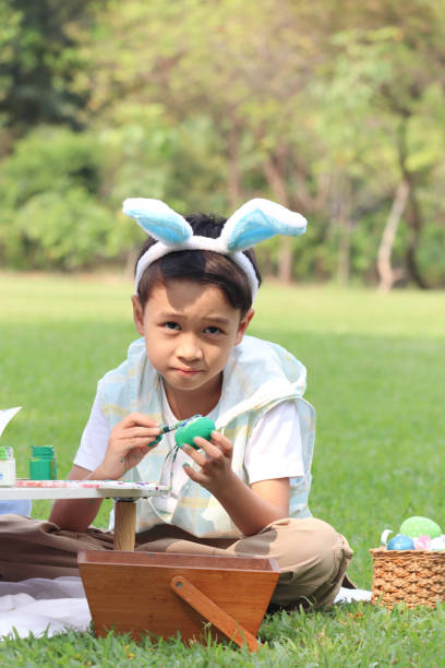 ウサギの耳を持つかわいいアジアの男の子は、自然の庭の緑の草原に座って絵筆で卵を描いています。イースター休暇を屋外で祝う子供。幸せな子供はイースターに公園で楽しんでいます - easter easter bunny fun humor ストックフォトと画像