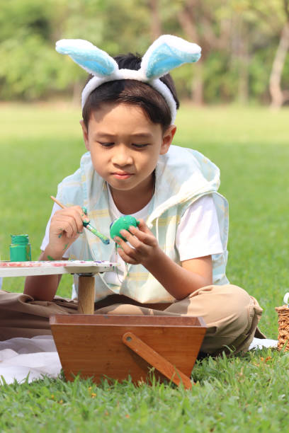 cute asian boy with bunny ears painting eggs with paintbrush while sitting on green grass meadow in nature garden. kid celebrating easter holiday outdoor. happy child has fun in park on easter - easter easter bunny fun humor ストックフォトと画像