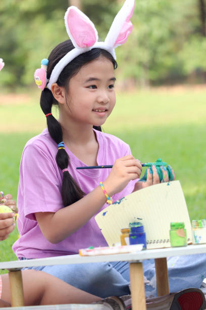 ウサギの耳を持つかわいいアジアの女の子は、自然の庭の緑の草原に座っている間、絵筆で卵を描いています。イースター休暇を屋外で祝う子供。幸せな子供はイースターに公園で楽しんで� - easter easter bunny fun humor ストックフォトと画像