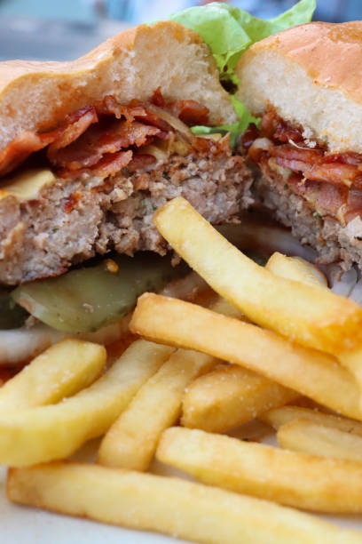 istock Full frame image of bacon cheeseburger in white bread burger bun, beef patty, melted cheese slice, onion rings and gherkin slices served with pile of crispy French fries, lettuce side salad, wooden table background, focus on foreground 2063336155