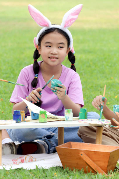 ウサギの耳を持つかわいいアジアの女の子は、自然の庭の緑の草原に座っている間、絵筆で卵を描いています。イースター休暇を屋外で祝う子供。幸せな子供はイースターに公園で楽しんで� - easter easter bunny fun humor ストックフォトと画像