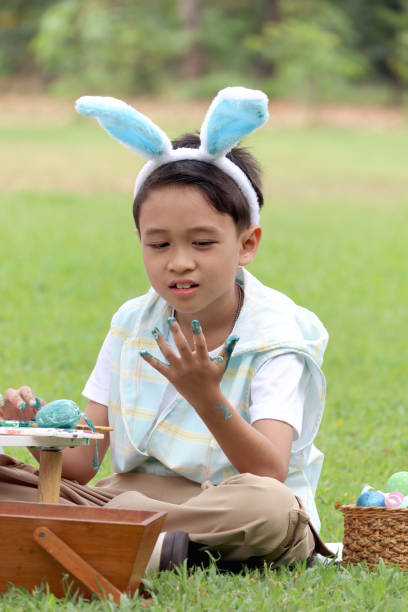 自然の庭の緑の草原にブラシで卵を描いた後、手を見ている青いウサギの耳を持つかわいいアジアの男の子。イースター休暇を屋外で祝う子供。幸せな子供は公園で楽しんでいます。 - easter easter bunny fun humor ストックフォトと画像