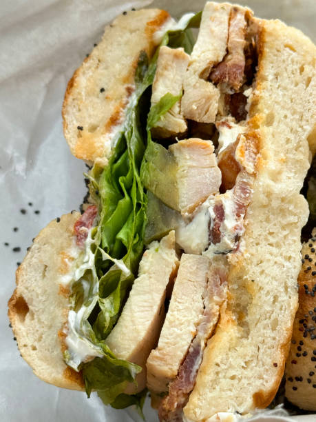 istock Full frame image of halved fresh, sesame seed bagel filled with chicken, bacon, lettuce and mayonnaise, greaseproof paper wrapping, elevated view 2063346404