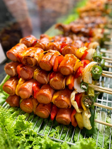 Close-up image of grilled frankfurter sausage kebabs on bamboo skewers with red and yellow bell peppers, onion and green chillies, lying in rows on metal grill rack over banana leaves, cooked sausages with sticky spiced sauce, focus on foreground