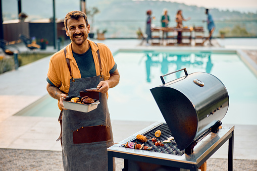 Happy man making barbecue while having party with friends by the pool and looking at camera. Copy space.