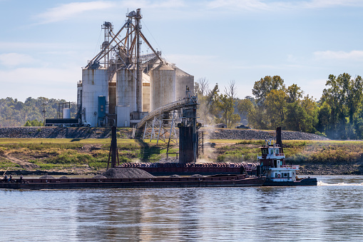 Tugboat pushing freight barges past grain loading dock in farming country in Kentucky