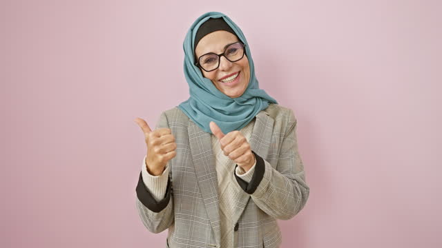 Smiling, confident middle age hispanic woman in hijab pointing behind with thumbs up over isolated pink background