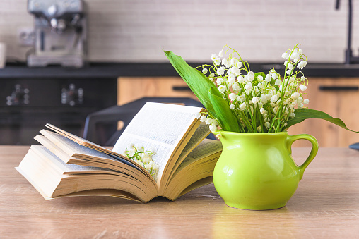 bouquet of lilies of the valley in green jug, open book on a wooden table in a kitchen