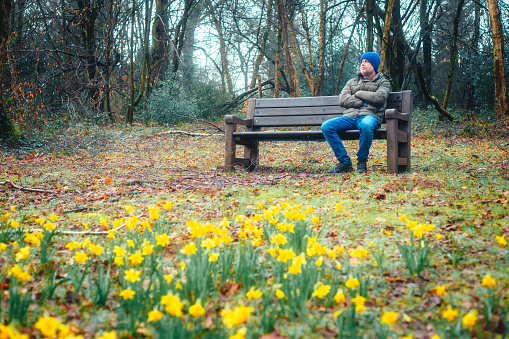 Color image depicting a sad, lonely mid-adult man sitting on a bench in the park on a cold morning in early Spring. He is wearing warm clothing and a blue knit hat.