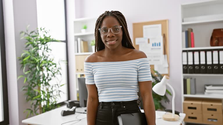 A confident young woman with braids and glasses stands in a modern office, exuding professionalism and poise.