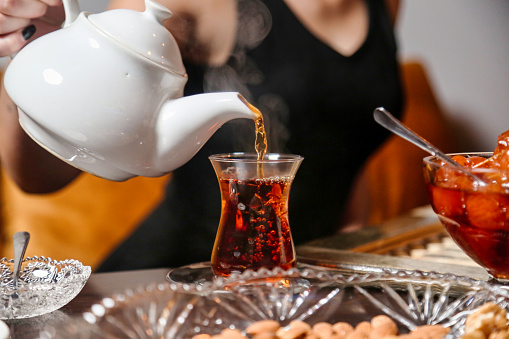 A woman carefully pours tea from a kettle into a glass teapot.