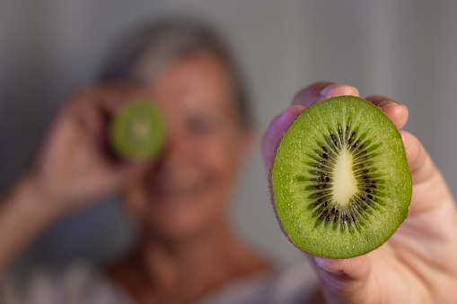 Defocused senior woman holding a ripe green kiwi cut in half, focus on kiwi. Healthy eating concept