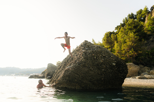Photo of a senior man looking at his grandson while he's jumping into the sea from the rock