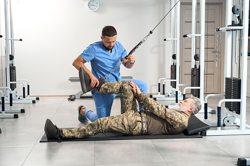 Physiotherapist helps a military man perform exercises to stretch his leg muscles on a special machine
