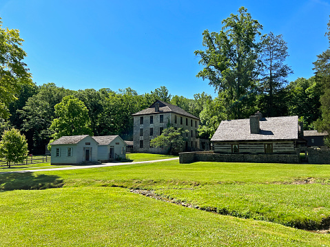 Mitchell, Indiana, USA - June 20, 2022:  Historic Grist Mill, Apothecary, Mercantile and Lower Residence exterior in the recreated and restored 1800 Pioneer Village at Spring Mill State Park, near Mitchell, Indiana with beautiful blue sky and vivid green trees and grass copy space.