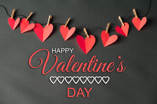 Red paper cut hearts hanging on a clothesline with wooden clothespins on a gray background. HAPPY VALENTINE'S DAY lettering is in the center. Can be used as a design for Valentine's day holiday greeting cards or posters.