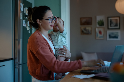 Mother, baby and kitchen with laptop at night for remote work, deadline or multi tasking at home. Mom or freelancer holding toddler or newborn while working late on computer at desk, table or house