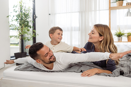 Photo of a lovely young family playing in bed. A father carries his cute son on his back and imitates a plane flying.