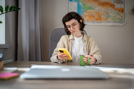 Serious teenage girl having tea break after freelance work, studying on distant education. Pensive teen sitting at table with cup of hot beverage, holding smartphone, scrolling web pages in internet