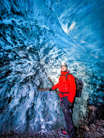 Man with a red jacket standing in front of the wall of a glacier in Iceland. Shot from inside the cave, the man is looking at the camera. Cave under the Vatnajokull glacier with a blue translucent ceiling