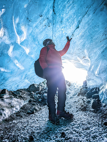 Man with a red jacket standing in front of the entrance of a glacier cave in Iceland. Shot from inside the cave, the man is looking up at the ice. Cave under the Vatnajokull glacier with a blue translucent ceiling