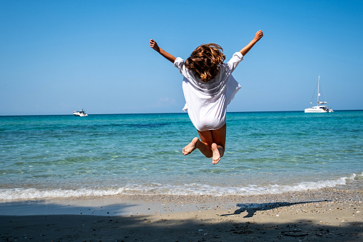 happy teenage girl with her back turned jumping and looking at the sea with open arms in a white shirt with curly hair