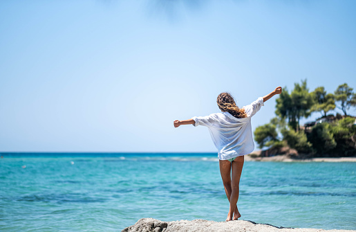 teenage girl on a rock at the sea with open arms looking at the sea in a white shirt and swimsuit, sunny day