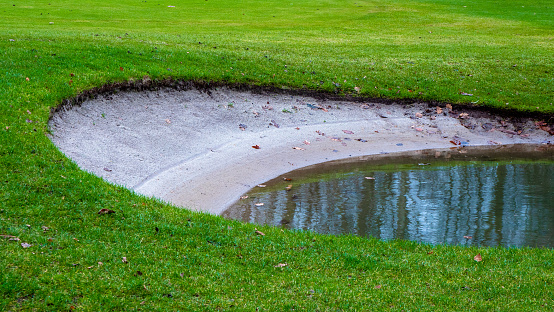 Closeup of a bunker in a neatly manicured golf course filled with rain water after a flood reflecting bare winter trees