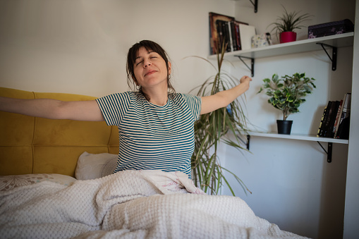 Young woman is woke up. She is sitting on the bed in her bedroom, covered with white blanket and she is  stretching. Starting a new day.