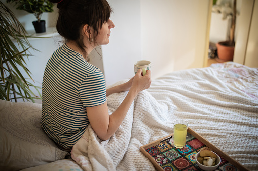 The side view of young woman in casual clothes with messy bun who is sitting on the bed, covered with white blanket and holding cup of coffee.Trey with fruits and juice is on the bed next to her. Beautiful morning in bedroom full of natural light.