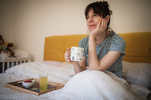 Young smiling woman with messy bun is sitting on the bed and holding cup of coffee. Waking up, smiling face and smell of coffee. Tray with juice and fruits is on the bed, next to her. The beginning of new day.