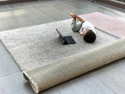 A young child is sprawled out on a large, textured gray carpet, comfortably using a laptop. The warmth of the indoor lighting suggests a casual, domestic setting during the day. The boy seems focused on the screen, potentially engaging in online learning or entertainment.