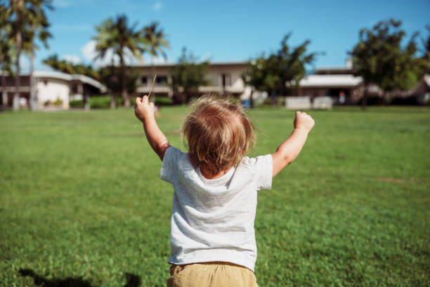 Rear view of a one year old boy standing in the grass with his arms raised in celebration on a beautiful and sunny day in Hawaii.