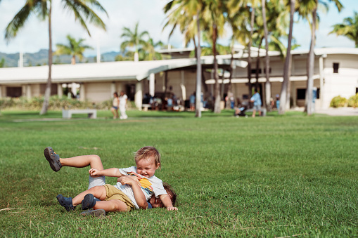 A cute one year old Eurasian boy giggles while wrestling with his sister in the grass at a park in Hawaii.